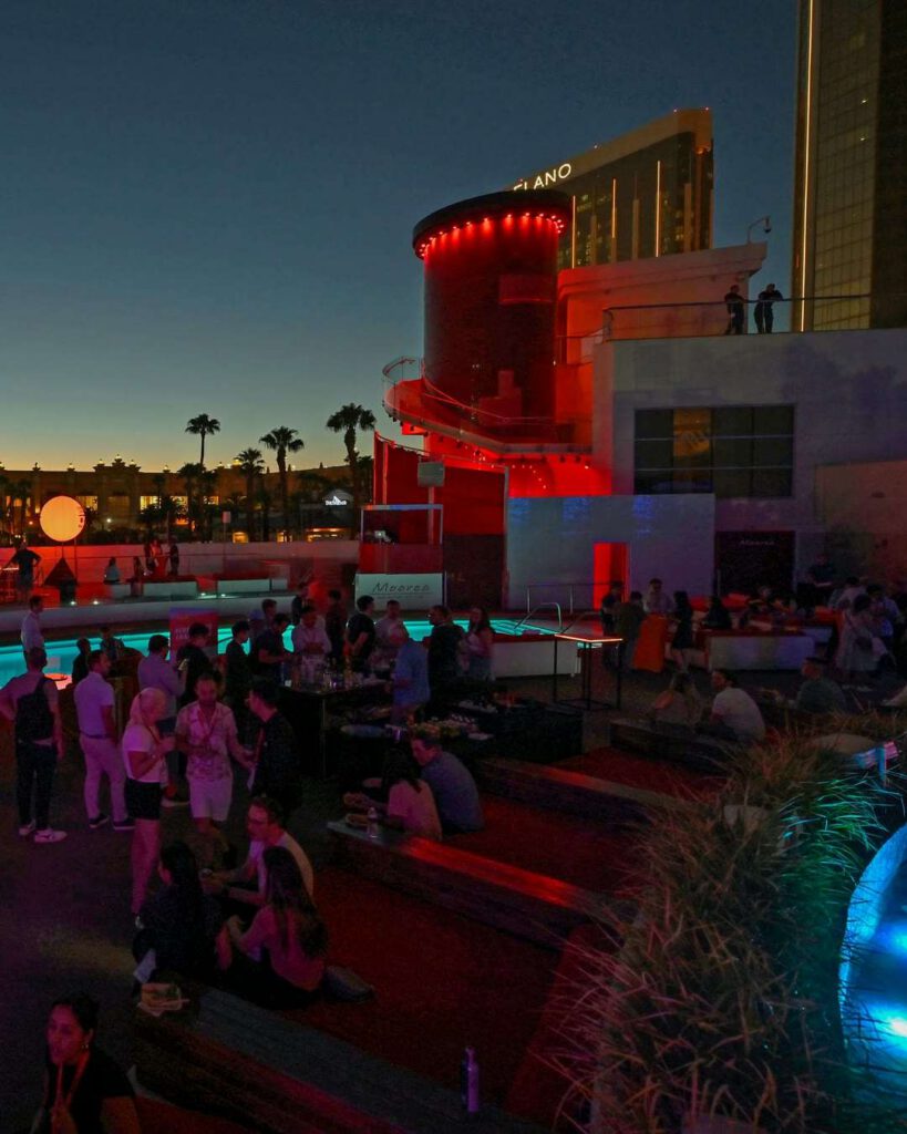 Soirée au bord d'une piscine au crépuscule, tour cylindrique éclairée de rouge, palmiers en silhouette et foule animée