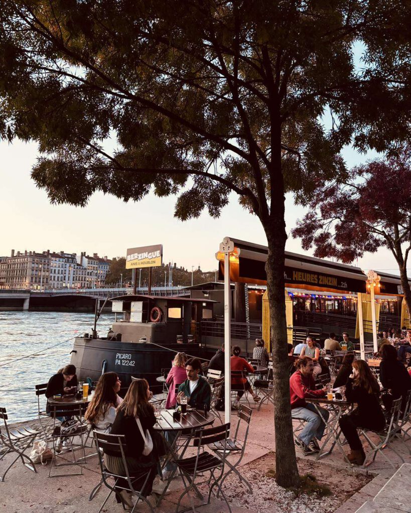 Terrasse de la guinguette Le Berzingue au bord de la Saône au crépuscule, péniche-bar et tables animées sous un arbre