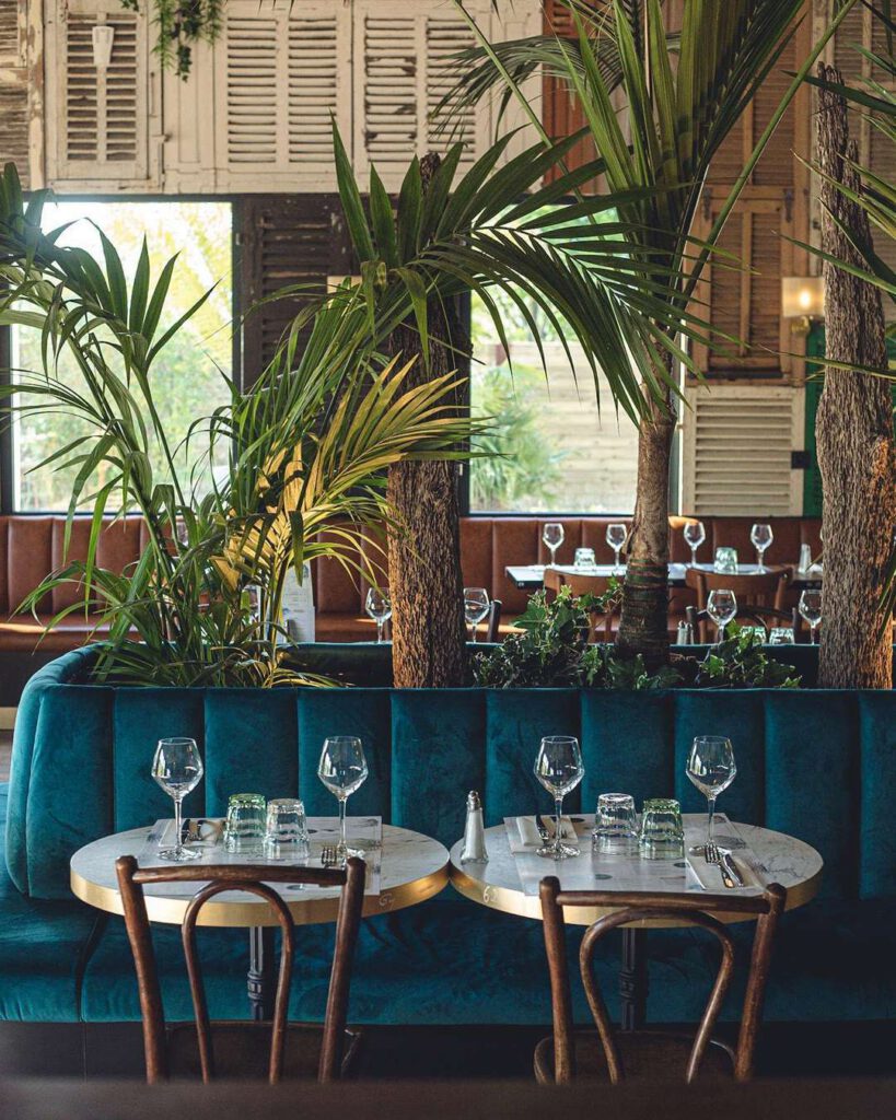 Intérieur tamisé d'un bistrot : banquette en velours bleu, tables rondes en marbre dressées, palmiers et volets en bois