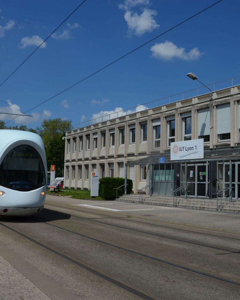Façade de l'IUT Lyon 1 par une journée ensoleillée, tramway bleu à l'arrêt le long de la voie