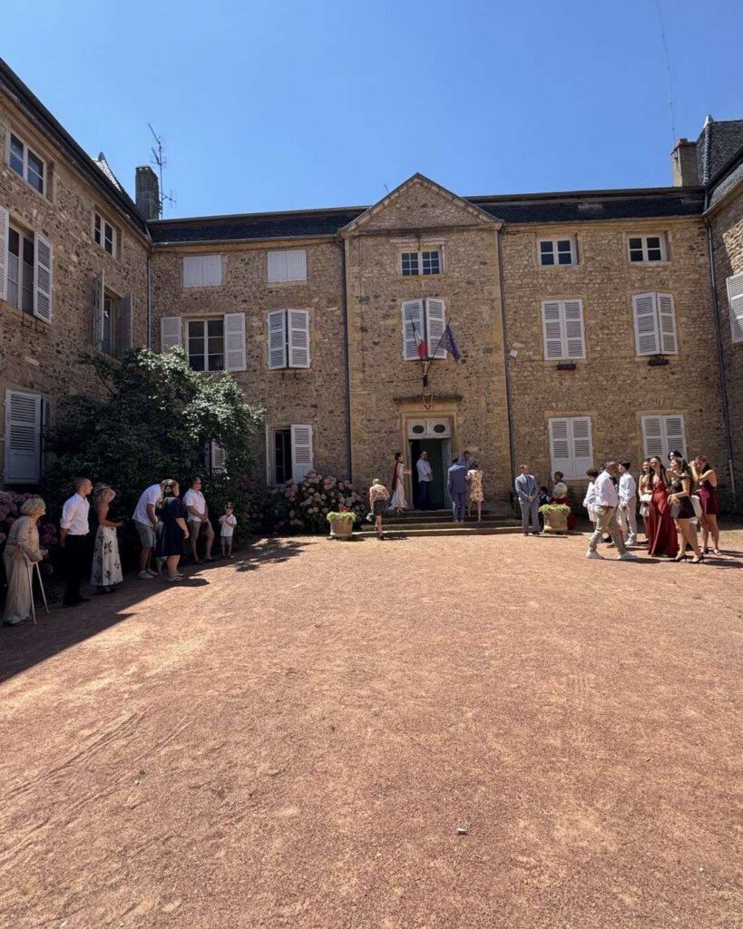 Mariage dans la cour d'un château en pierres dorées, invités rassemblés devant l'entrée pavoisée