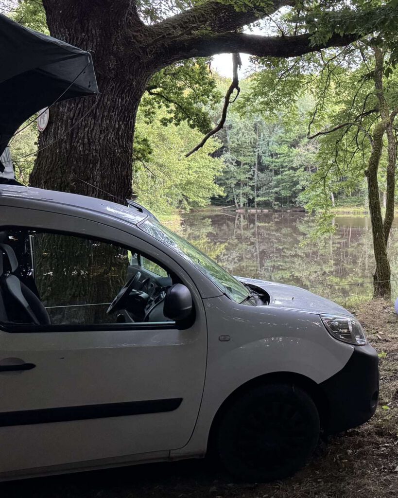 Campement au bord d'un étang en forêt : van blanc avec tente de toit ouverte sous un grand chêne, eau calme en arrière-plan