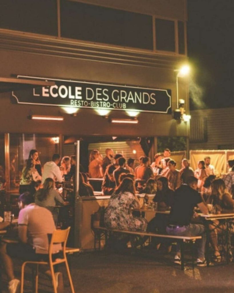 Terrasse animée du bar-club L'École des Grands de nuit, enseigne éclairée et tablées de convives