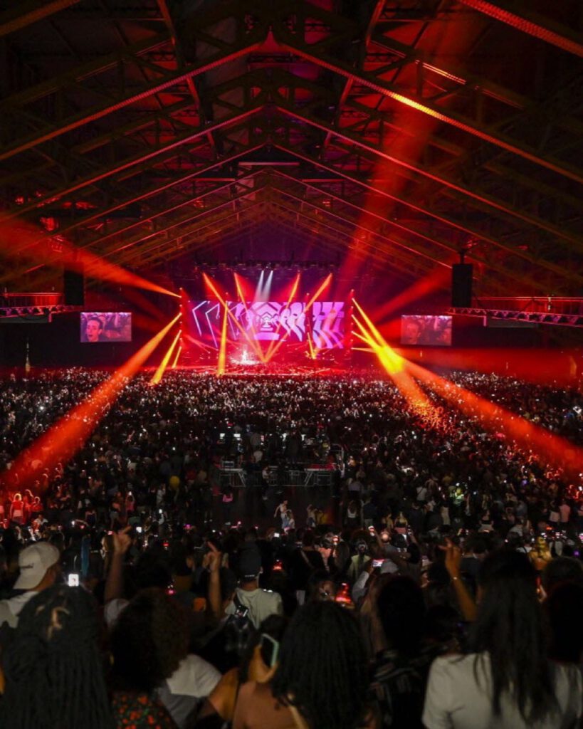 Concert dans une grande halle : charpente rouge, faisceaux lumineux convergents et foule immense face à la scène
