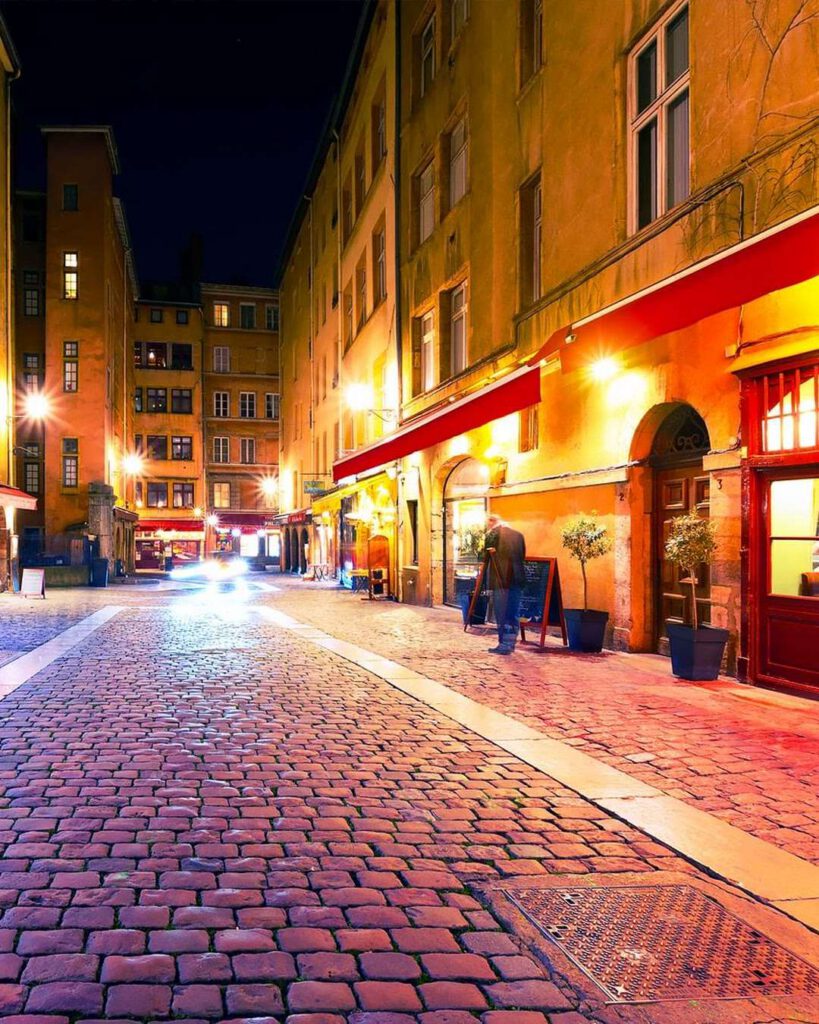 Rue pavée du Vieux Lyon la nuit, façades chaleureuses éclairées et devantures de restaurants aux stores rouges