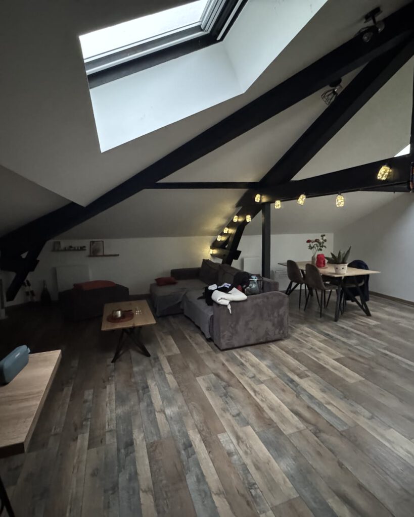 Appartement mansardé sous les toits : velux, poutres noires apparentes, guirlande lumineuse, canapé et table dressée sur un parquet bois.