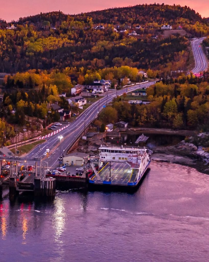 Traversier de Tadoussac à quai au crépuscule, route descendant entre des collines aux couleurs d'automne sous un ciel rose.