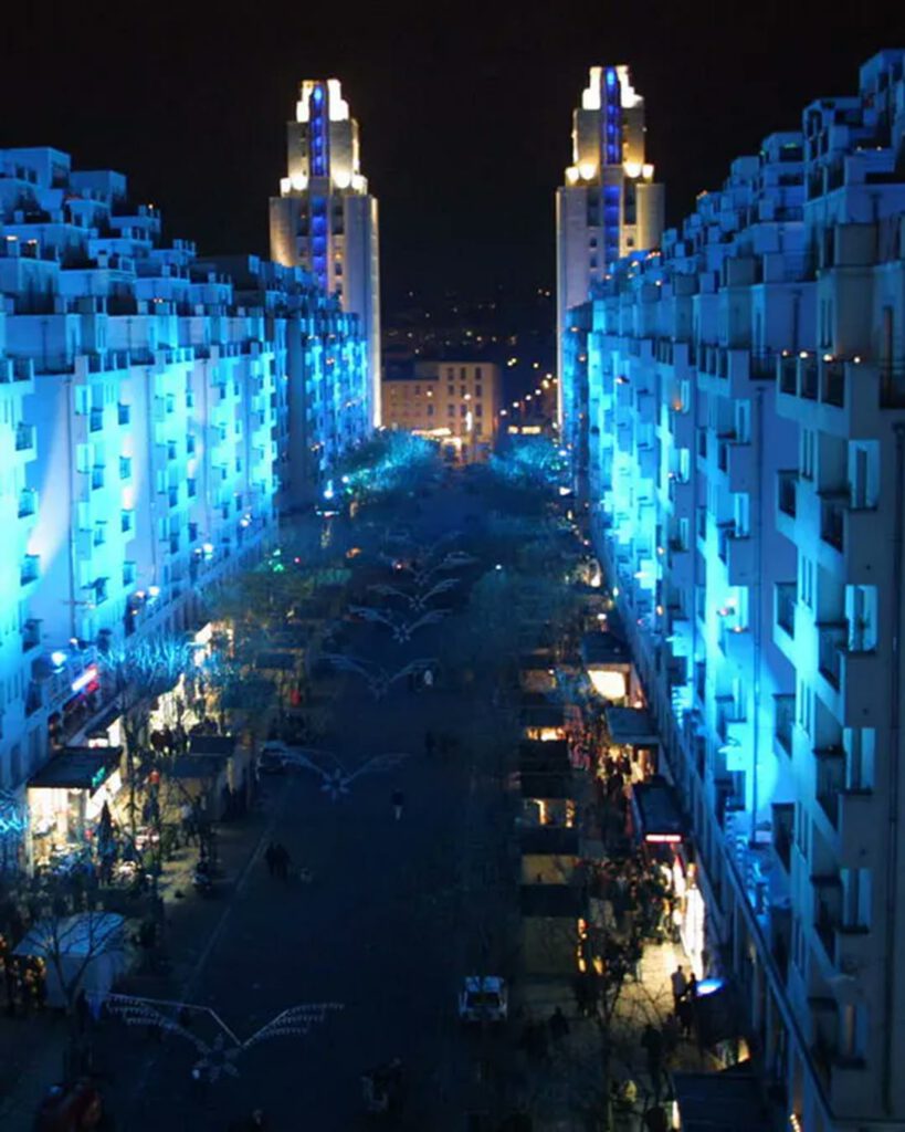 Avenue nocturne bleutée des Gratte-Ciel de Villeurbanne, tours jumelles illuminées et marché de nuit au pied des immeubles.