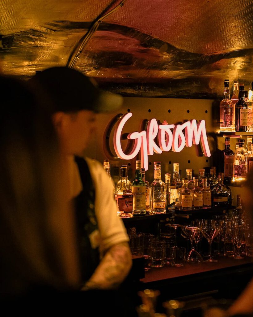 Intérieur tamisé du bar Le Groom à Lyon : néon rose « Groom », étagères de bouteilles et barman en contre-jour.