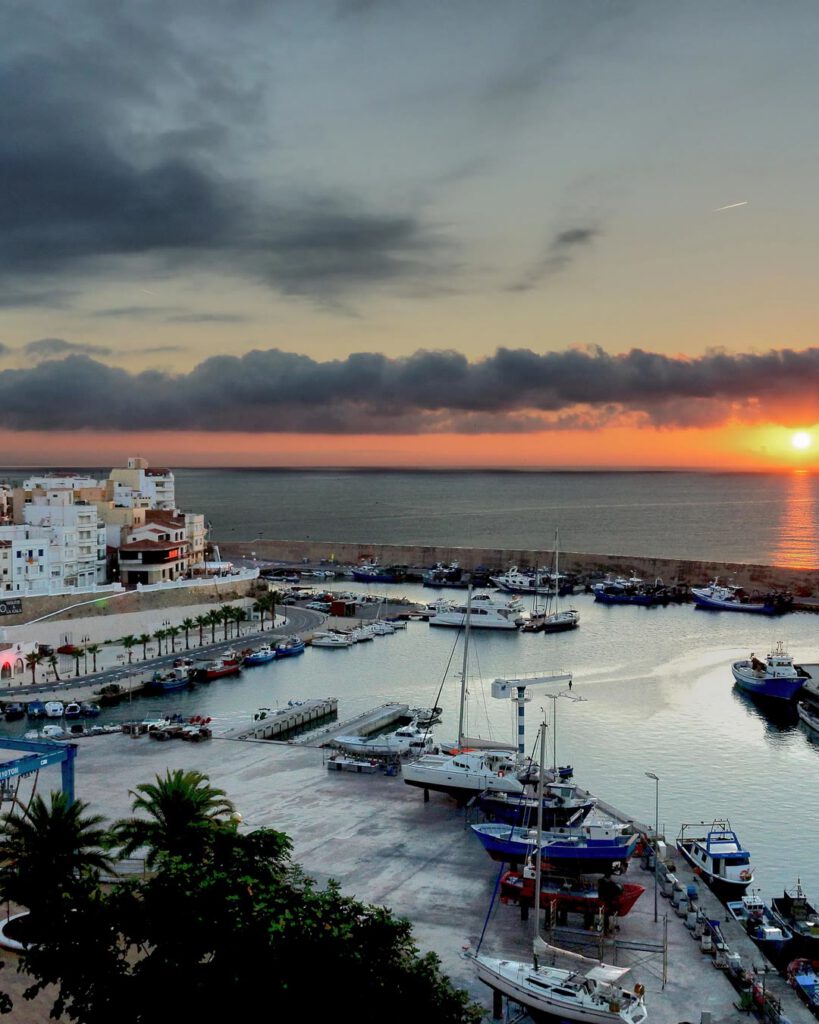 Port méditerranéen au coucher de soleil : bateaux amarrés, ville blanche à flanc de colline et soleil orange posé sur la mer.