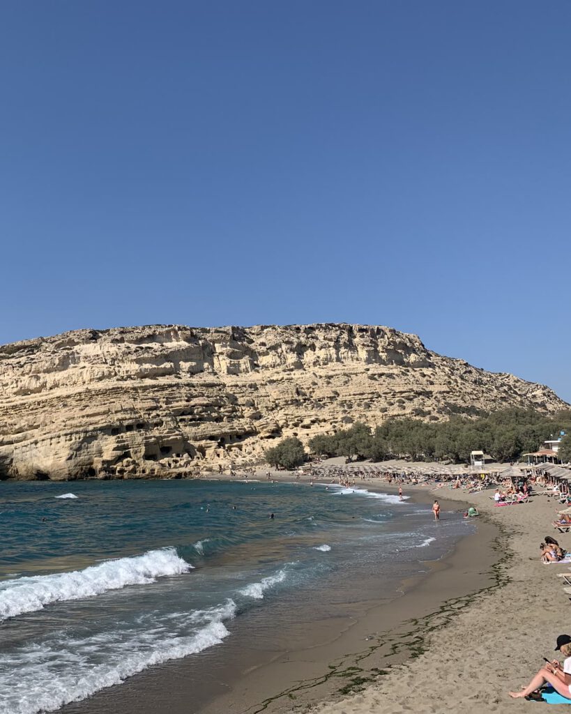 Plage de Matala en Crète : haute falaise percée de grottes, mer turquoise et bande de sable bordée de parasols.
