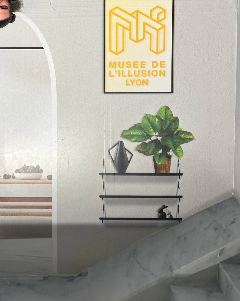 Entrée du Musée de l'Illusion de Lyon : affiche jaune et illusion d'optique d'une personne allongée au plafond, plante et étagères.