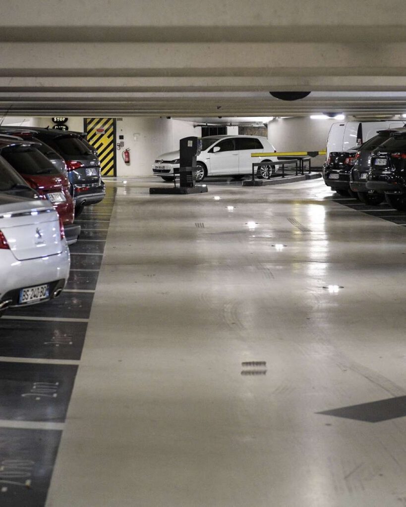 Parking souterrain faiblement éclairé : allée centrale, voitures alignées et porte de secours rayée jaune et noir