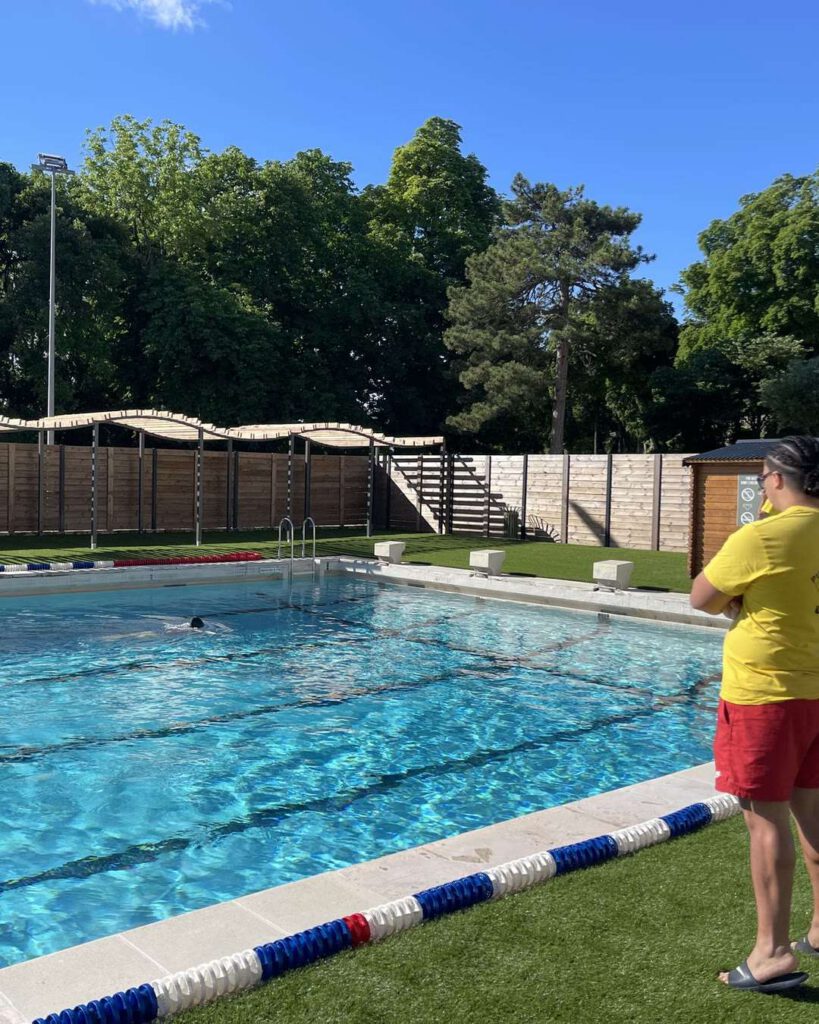 Piscine extérieure ensoleillée bordée d'arbres, lignes d'eau bleues et maître-nageur en tenue jaune et rouge au bord