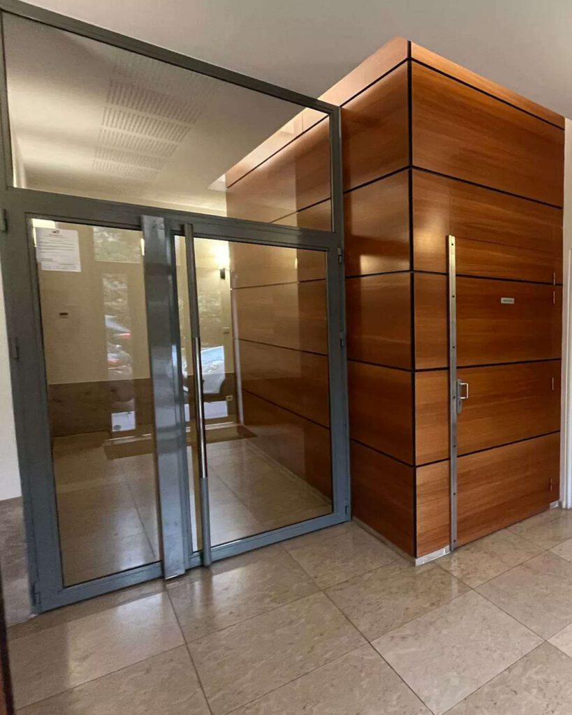 Hall d'immeuble cossu : porte d'entrée vitrée à cadre gris, mur d'ascenseur en panneaux de bois et sol carrelé